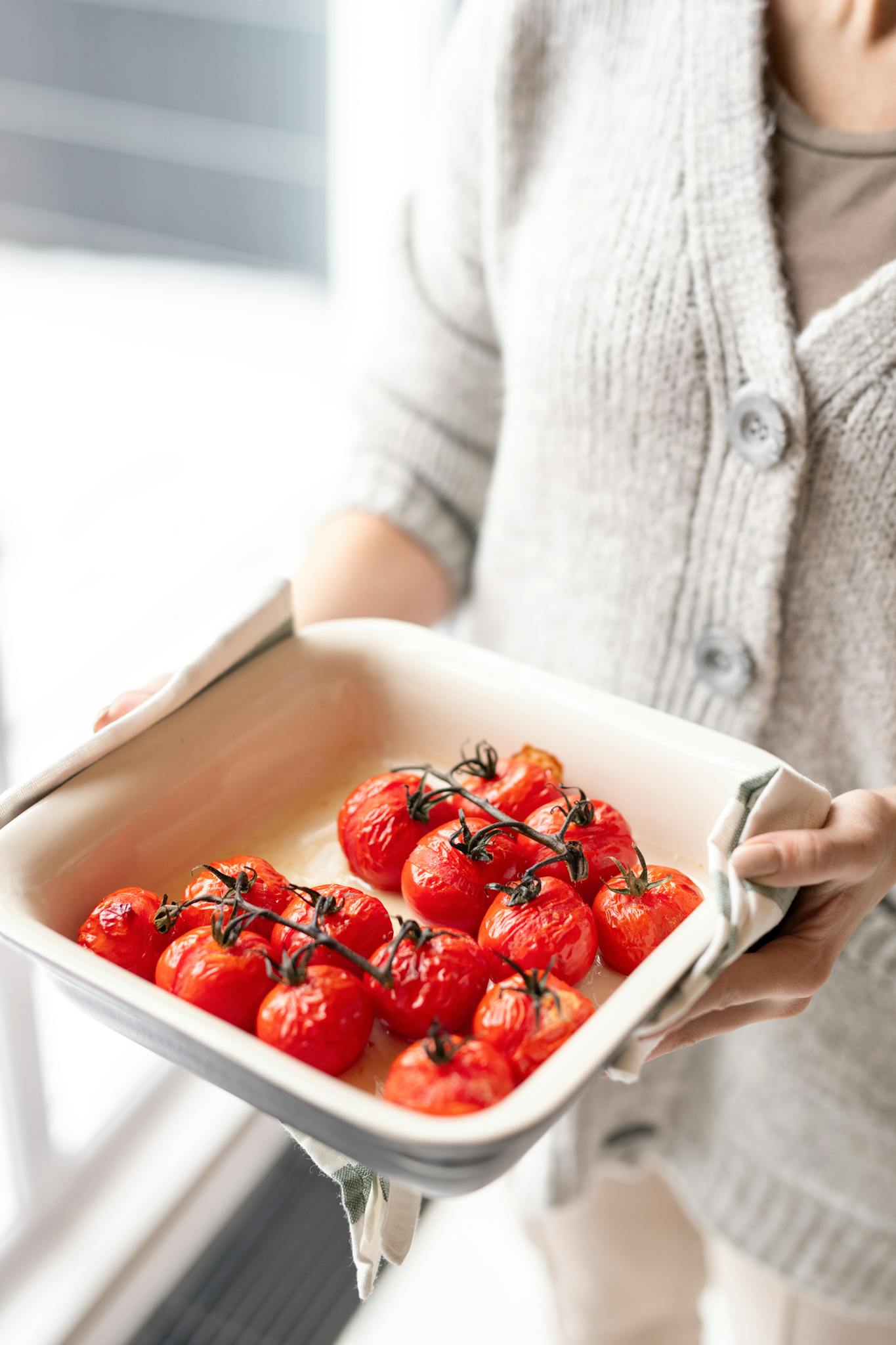 A woman in a cozy sweater holds a tray of freshly roasted red cherry tomatoes on the vine.